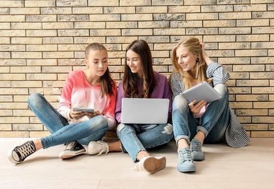 1131789908 Symbolbild Laptopversicherung. Bild zeigt drei Mädchen sitzend am Boden einer Schule. Alle haben Laptop oder Tablet in der Hand.