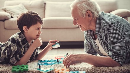 642510826 Opa und Enkel spielen im Wohnzimmer und lachen sich gegenseitig an.