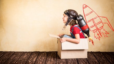 Kind mit Pilotenmütze und einer Raketennachbildung am Rücken sitzt in einem Karton